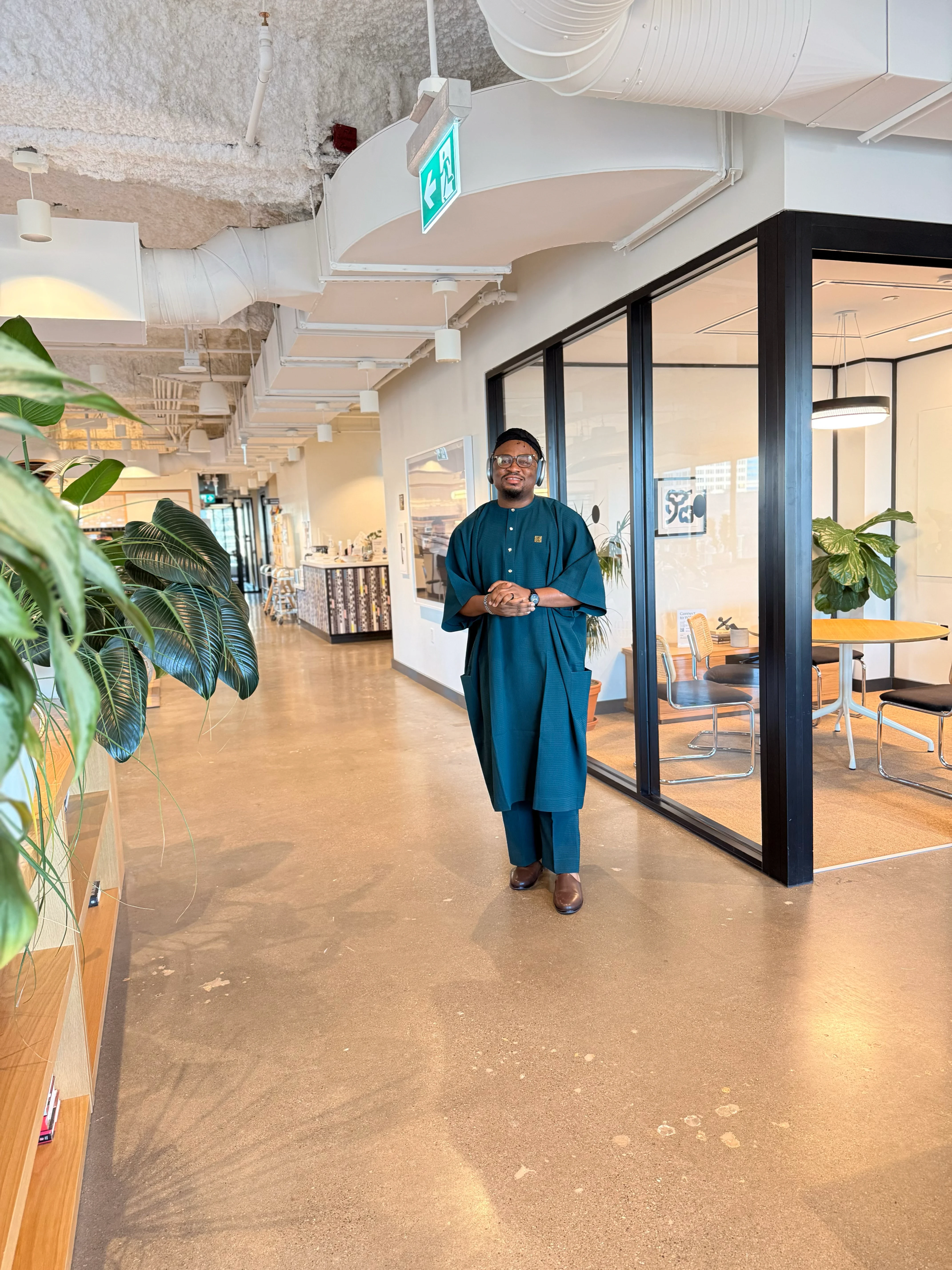 Oluwayemi in a teal agbada, standing in an office corridor beside a glass meeting room and a wall of plants